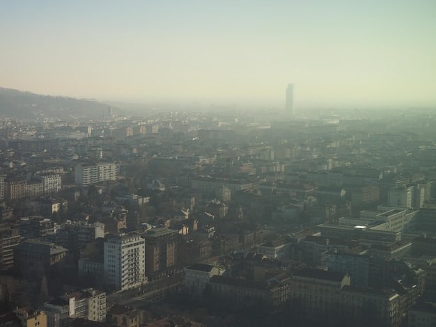 A smoggy cityscape seen from a distance, with a thick layer of air pollution obscuring the tops of buildings. The sky is a hazy gray color, emphasizing the negative environmental impact of traffic and industrial emissions.