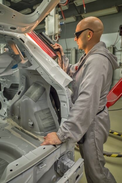 A photograph of a partially assembled car on a production line, with a worker holding a semiconductor chip in the foreground. The image symbolizes the direct impact of the chip shortage on the automotive industry and the challenges faced in maintaining production.