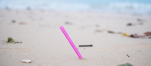 A close-up shot of a single plastic straw discarded on a beach, surrounded by sand and other plastic debris. The focus is on the straw, highlighting its role in marine pollution and the broader issue of 